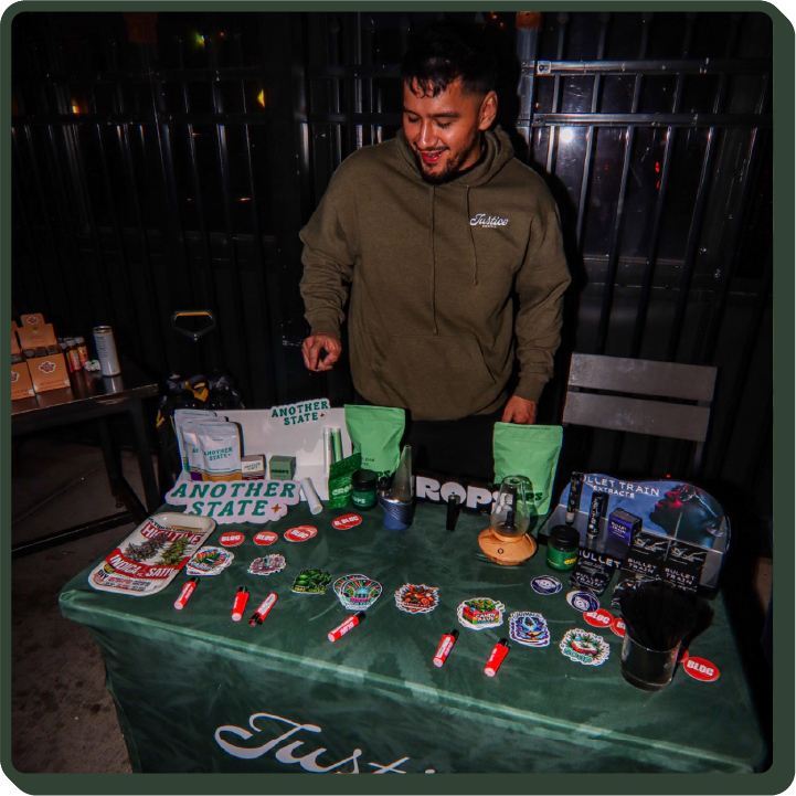 Man wearing a justice grown hoodie standing behind a display of Justice Grown products. There is an array set out of stickers, lighters, and products from our brands (Another State, Crops, Bullet Train, Bloc)